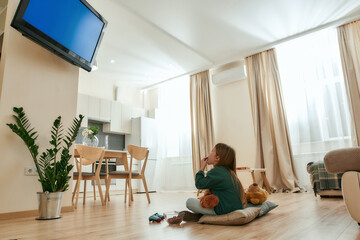 A cute little girl sitting infront of a TV on the floor eating cereal balls holding a teddybear in a spacy room