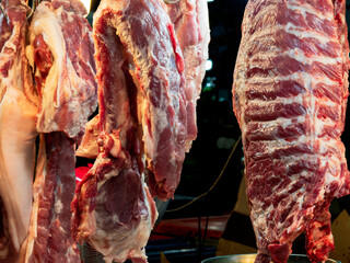 Pork ribs, large chunks of pork ribs (focus), and shrouded (blurred) pork are hung on the bars of a market pork stall.