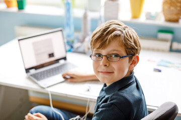 Kid boy with glasses learning at home on laptop for school. Adorable child making homework and using notebook and modern gadgets. Home schooling concept.