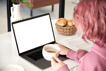 Hipster teen girl young woman student, worker sitting at kitchen table using laptop mock up white screen drink coffee learning online, working from home office, paying bills in app. Over shoulder view