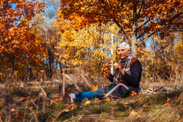 Naklejka na ściany i meble Senior alone man sitting in park picking yellow leaves. Retired man relaxing outdoors enjoying autumn nature