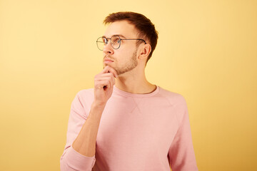 Portrait of a guy wearing glasses with a pensive face, holding a chick and thinking, dressed in pink casual isolated on yellow background