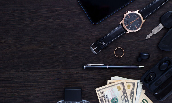 Men's Accessories On A Dark Wooden Background . The Concept Of Success And Business. Wrist Watch, Pen, Perfume, Headphones, Ring, Car Keys, Money And Phone. Flatlay. Copy Space.