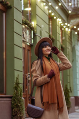 Outdoor fashion, lifestyle portrait of happy smiling woman wearing elegant autumn, winter outfit posing in street of European city. Model dressed trendy beige coat, hat, brown scarf, with stylish bag