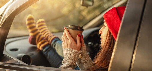Woman in a car in warm socks yellow is holding a Cup of coffee in hands at sunset. Cozy autumn weekend trip