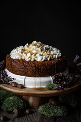 Homemade Carrot cake with ricotta cheese and honey frosting on wooden cake stand surrounded with moss, acorns, pines and with little white candle on foreground on black background.