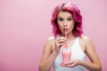 surprised young woman with colorful hair drinking strawberry milkshake from straw isolated on pink