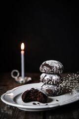 Homemade chocolate cookies  in pile on white plate and lit candle on old wooden table on dark background .