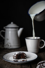 Homemade chocolate cookies on white plates and pouring milk into white and blue cup on old wooden table on dark background .