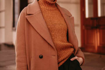 Young woman  with cropped head in the orange knitted cozy sweater and brown coat standing on the...