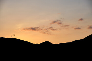 A fiery sky is lit up by the late sunset tones on a summers evening on the Isle of Mull, Scotland. Silhouetted mountains are visible and two wind turbines sit on the tops.