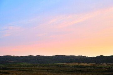 Naklejka na ściany i meble A pastel coloured sky overlooks beautiful fields of cows and sheep during a beautiful sunset on the Isle of Mull, Scotland in summer. Blue and pink and yellow colours light up the sky.
