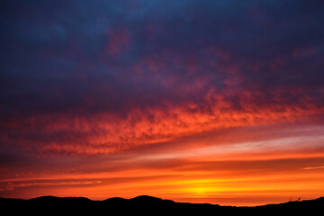 A fiery sky is lit up by the late sunset tones on a summers evening on the Isle of Mull, Scotland. Silhouetted mountains are visible at the bottom of the image and orange and reds light up the sky.