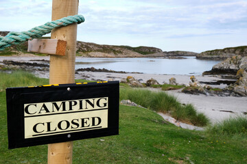A temporary Camping Closed sign is stuck to a post in front of the beautiful Uisken Beach on the Isle of Mull, Scotland. Green grass up to the waters edge and seaweed on the sandy beach.