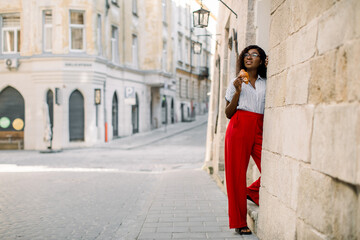 Fototapeta na wymiar Charming young African woman in red pants, posing to camera with fresh tasty croissant, standing outdoors on the background of old vintage city buildings.