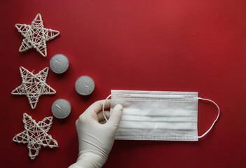 female gloved hands holding medical mask with Christmas decorations on a red background. Copyspace. Flatlay