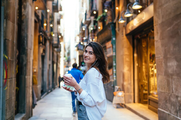 Half length portrait of carefree hipster girl with takeaway caffeine beverage and smartphone in hands smiling at camera during touristic travelling, pretty woman in earphones enjoying music playlist