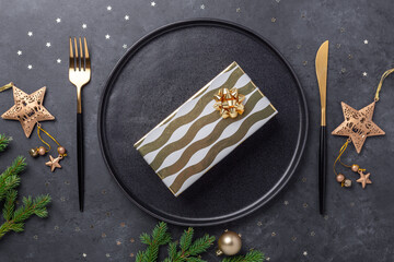Christmas table setting with black ceramic plate, gift box and gold accessories on black stone background. Top view. Copy space