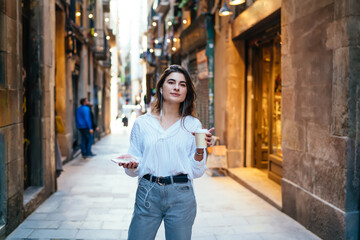 Thoughtful hipster girl in electronic earphones looking away while waiting guide for travel sightseeing, Caucasian female tourist with coffee to go and cellular technology listening online book