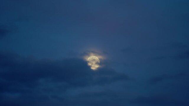 Full moon and clouds are slow moving through each other in dark blue sky. Moonrise in twilight.
