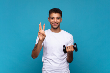 young man lifting a sumbbell smiling and looking friendly, showing number three or third with hand...