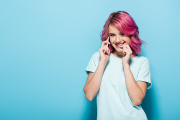 young woman with pink hair talking on smartphone and smiling on blue background