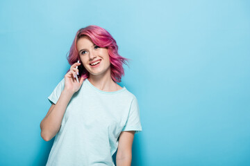 young woman with pink hair talking on smartphone and smiling on blue background