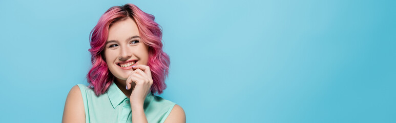 young woman with pink hair smiling and flirting isolated on blue, panoramic shot