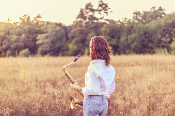 Young red-haired woman in a white shirt with a tenor saxophone in her hands stands in an autumn field and looks at the sunset - obrazy, fototapety, plakaty
