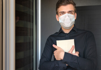 Portrait of a young man in a protective medical mask, at the open door, with a book in his hands. The concept of quarantine, online training, and the  second wave of coronavirus.