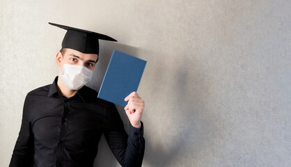Portrait of a young student in a graduation cap and protective mask with a diploma in his hand, on a gray background, banner. The concept of distance education in the context of the coronavirus pandem