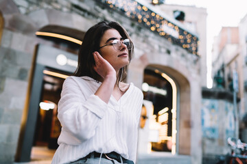 Pondering hipster girl in classic spectacles for provide eyes correction and electronic headphones listening recorded sounds while resting in city, thoughtful female enjoying pastime with music hobby