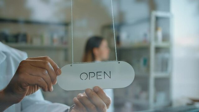 Unrecognizable man turning the plate over from closed to open on pharmacy door. Blurred form of woman placing medications in the bg. Reflection of a street traffic and passing cars in a glass door.
