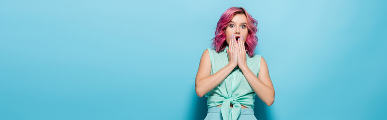 shocked young woman with pink hair covering mouth on blue background, panoramic shot