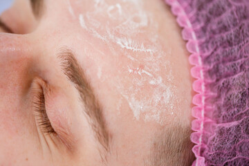Portrait of young man on cleaning face procedure by woman cosmetologist in beauty clinic. Beutician doctor is wiping patient's face with cotton pads. Beauty industry concept. Guy is lying on couch.