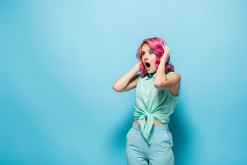 shocked young woman with pink hair and open mouth holding head on blue background