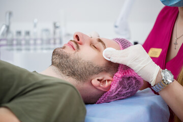 Portrait of young man on cleaning face procedure by woman cosmetologist in beauty clinic. Beutician doctor is wiping patient's face with cotton pads. Beauty industry concept. Guy is lying on couch.
