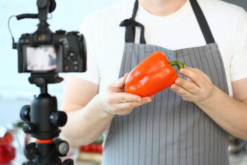 Chef Vlogger Showing Camcorder Orange Pepper. Man in Apron Recording Ripe Vegetable on Camera for Culinary Vlog in Kitchen at Home. Male Hands Holding Fresh Ingredient Horizontal Photography