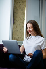 A young woman is sitting on the couch and working on a laptop remotely from work. Girl with a computer on her knees looking at the monitor