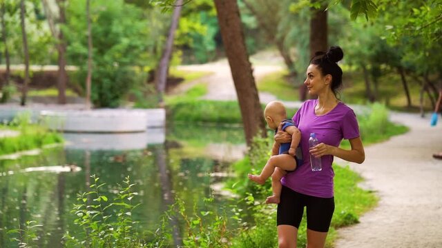 Young woman wearing sportswear carrying baby and drinking water after training in park. Sporty mother and little child spending time together. Concept of fitness