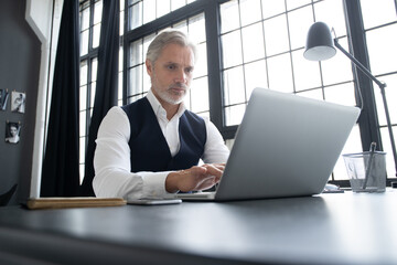 Concentrated at work. Mature man in full suit using laptop while working in modern office