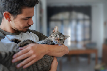 Hombre joven sostiene un gato atigrado en sus brazos y lo mira con amor