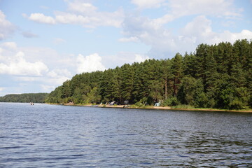 Scenery forestry riverbank with camp site, view from water Volga river on blue sky with white clouds background, beautiful river landscape at summer day