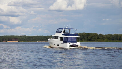 Modern white flybridge motor yacht with inboard motor slowly floating on calm water on forest river shore and blue cloudy sky background, side rear view at summer day, powerboat luxury recreation 