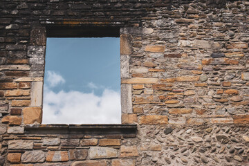 Window in ancient stone wall of damaged house, toned. Abandoned medieval building, filtered. Blue sky in window of ruin temple. Destroyed facade of old stone building. History concept. 