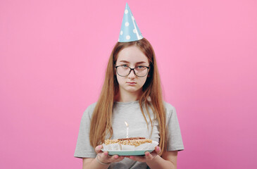 Front view of unhappy young lady wearing funny party hat holding birthday cake isolated on pink