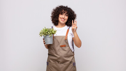 young woman gardener smiling and looking friendly, showing number three or third with hand forward,...