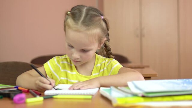 Little cute girl sits at a desk in the classroom. Draws in a notebook with a felt-tip pen. Preschool education. 4k