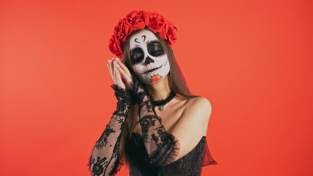 Day of Dead, lady with skull make-up putting face on folded hands and acting like sleeping, smiling posing on red background, Halloween
