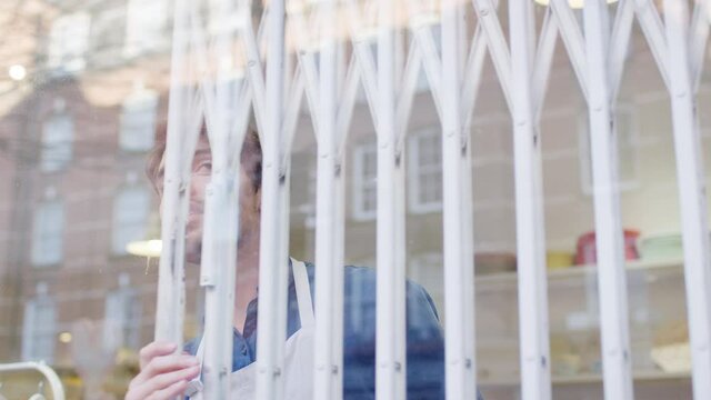 Man running small business pushing back security grill from window and opening shop viewed from outside - shot in slow motion